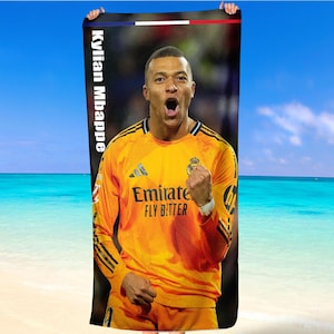 May include: A beach towel with a picture of Kylian Mbapp&eacute; in a yellow football jersey. The towel has the text "Kylian Mbappe" vertically on the left side. The background shows a beach and blue sky.