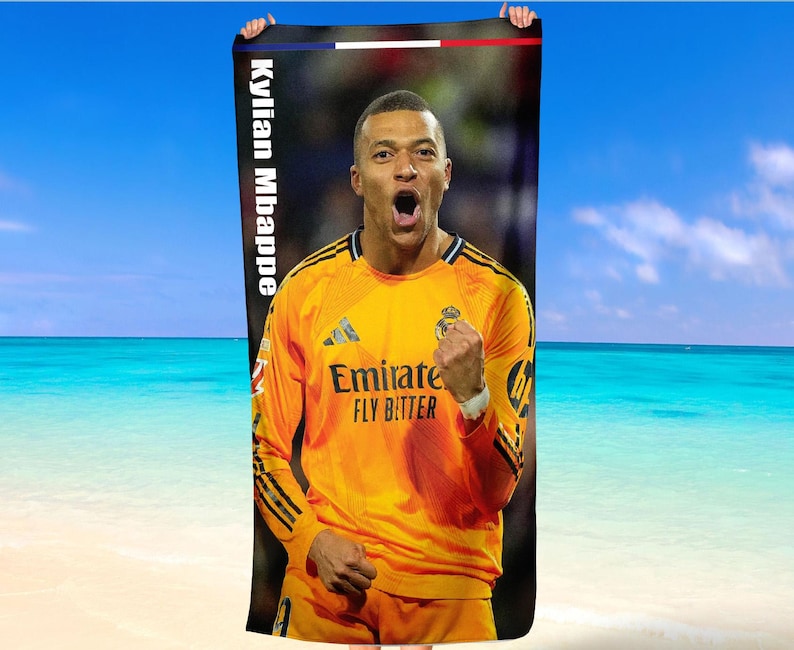 May include: A beach towel with a picture of Kylian Mbapp&eacute; in a yellow football jersey. The towel has the text "Kylian Mbappe" vertically on the left side. The background shows a beach and blue sky.
