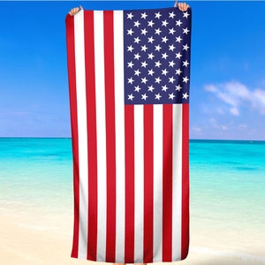 May include: A rectangular beach towel featuring the American flag design. The towel has red and white vertical stripes and a blue square in the upper left corner with white stars. The towel is held up against a backdrop of a beach and blue sky.