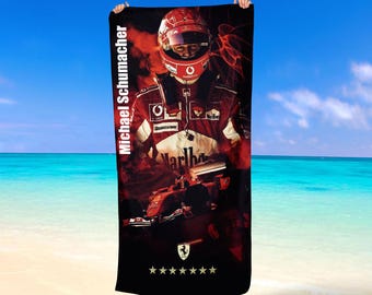 Può includere: Telo mare nero con un ritratto di Michael Schumacher con tuta e casco da corsa. Il telo ha la scritta "Michael Schumacher" in bianco, con un logo Ferrari e cinque stelle in basso. Lo sfondo presenta un design rosso e nero.