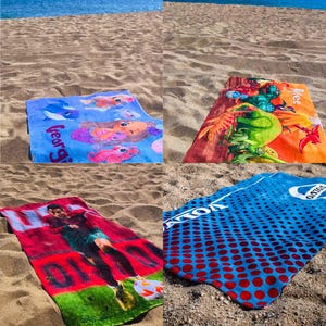May include: Four beach towels are laid out on sand. One is blue with a mermaid design and the name "Georgia". Another is orange with a cartoon character. A third towel features a soccer player. The last towel is blue with red dots.