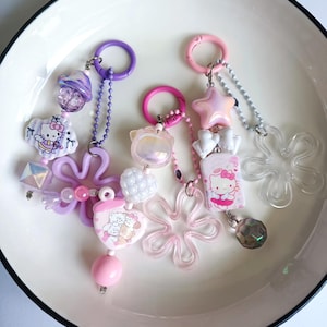 May include: A collection of colorful keychains with various charms, including Hello Kitty designs, stars, and flower shapes. The keychains feature pink, purple, and clear accents, with metal clasps and beaded chains. The keychains are displayed on a white plate.