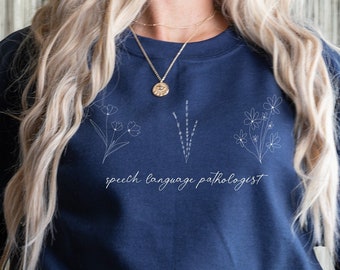 Puede incluir: Una sudadera azul marino con un diseño floral blanco y el texto "speech language pathologist".