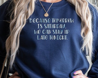 Puede incluir: Sudadera azul marino con texto blanco: "BECAUSE TOMORROW IS SATURDAY, WE CAN STAY UP LATE TONIGHT." La sudadera se usa con jeans azules. La persona tiene el pelo rubio.