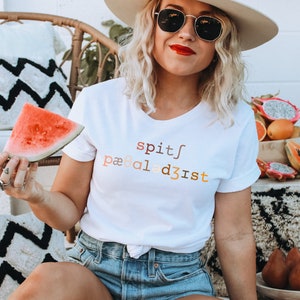 Op de afbeelding: Een wit T-shirt met de tekst "spit/paaled3ist" in een regenboogkleurige gradi&euml;nt. Het shirt wordt gedragen door iemand die een stuk watermeloen vasthoudt.