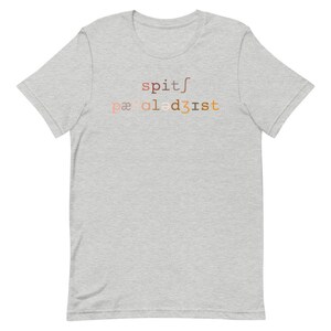 Op de afbeelding: Een grijs t-shirt met de tekst "spit/paaled3ist" gedrukt op de voorkant in een verloop van bruin, oranje en roze.