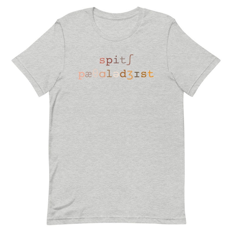 Op de afbeelding: Een grijs t-shirt met de tekst "spit/paaled3ist" gedrukt op de voorkant in een verloop van bruin, oranje en roze.