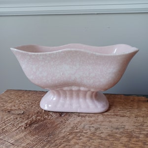 May include: A pale pink ceramic planter with a speckled white finish and a scalloped rim. The vessel features a flared, elongated body that tapers down to a ribbed, oval-shaped pedestal base, creating a distinct and balanced silhouette.