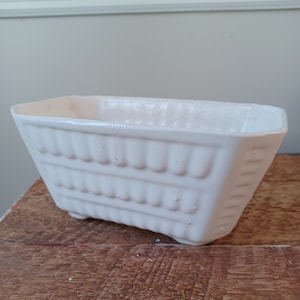 May include: A rectangular, off-white ceramic planter with a ribbed design. The planter has a slightly tapered shape and sits on small feet. The interior is smooth, and the exterior features a series of vertical ridges. The planter is on a wooden surface.