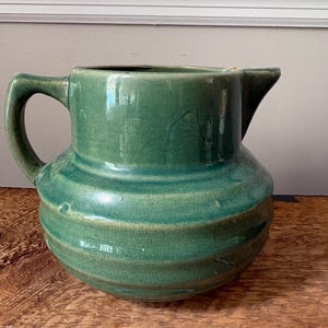 May include: A green ceramic pitcher with a glossy, crackled glaze finish. The vessel features a wide, rounded body with horizontal ribbed detailing, a sturdy side handle, and a prominent pouring spout at the top rim of the container.