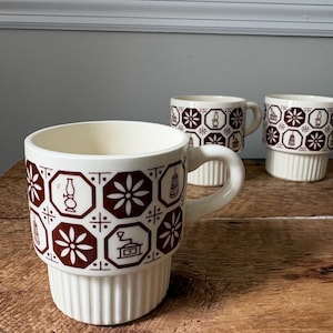 May include: Three cream-colored ceramic mugs featuring a repeating brown geometric pattern of octagons containing floral motifs, coffee grinders, and oil lamps. The mugs have a smooth upper section and a vertically ribbed base with a curved handle.
