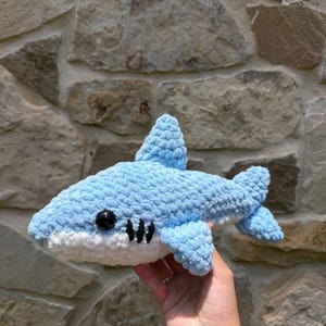 May include: A light blue and white crocheted shark plush toy with a textured yarn finish, featuring a prominent dorsal fin, a tail fin, and a black plastic eye. The shark has three black embroidered gill slits on its side and a white underbelly.