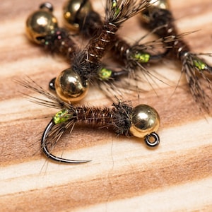 Può includere: Primo piano di diversi esche da pesca a mosca marroni e dorate con accenti verdi. Le esche sono legate con piume e hanno una testa di perline.