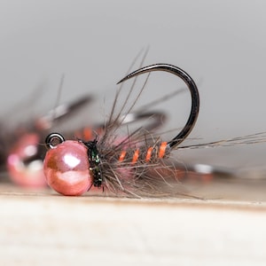 Può includere: Primo piano di un'esca da pesca con mosca con perline rosa, accenti arancioni e neri. L'esca ha un piccolo amo ed è legata con piume marroni.