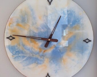 Puede incluir: Una esfera de reloj redonda, blanca, con remolinos abstractos azules y amarillos. El reloj tiene marcadores de hora en forma de diamante negro y manecillas de madera.