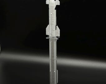 Può includere: Un modellino di razzo color argento posizionato verticalmente su una base circolare nera. Il razzo presenta un ogiva appuntita, due serie di alette laterali e un corpo cilindrico con dettagliate texture ad anelli orizzontali.