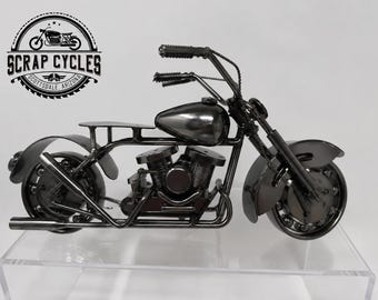 May include: A dark gray metal motorcycle sculpture. The motorcycle has a detailed engine, fenders, and handlebars. The sculpture is on a clear surface. The logo "SCRAP CYCLES SCOTTSDALE ARIZONA" is visible in the upper left corner.