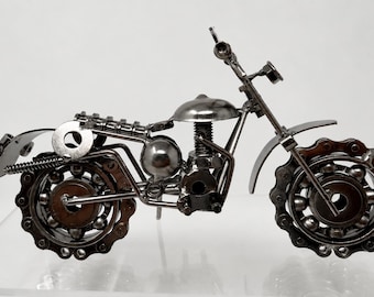 May include: A detailed metal sculpture of a motorcycle. The motorcycle features a silver fuel tank, handlebars, and fenders. The wheels are constructed from chains and ball bearings. The overall design is industrial and artistic.
