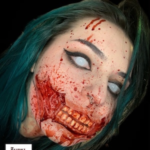 May include: Special effects makeup featuring a realistic prosthetic wound on the cheek exposing teeth and gums, covered in red blood splatter. The face has white mesh contact lenses, black eyeliner, and teal hair. Logo reads Trauma Queen FX.