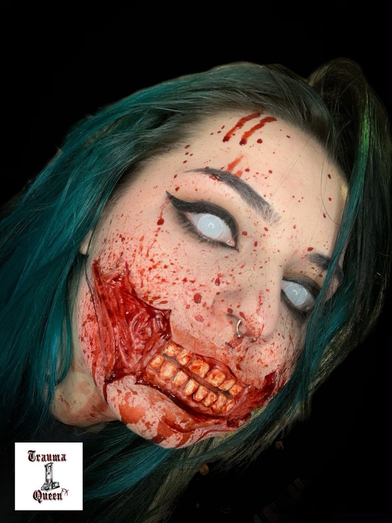 May include: Special effects makeup featuring a realistic prosthetic wound on the cheek exposing teeth and gums, covered in red blood splatter. The face has white mesh contact lenses, black eyeliner, and teal hair. Logo reads Trauma Queen FX.