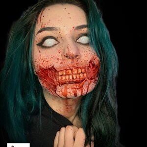 May include: A person with teal hair and white contact lenses featuring special effects makeup that simulates a torn mouth with exposed teeth and blood splatters across the face. A small logo in the corner displays the text Trauma Queen.
