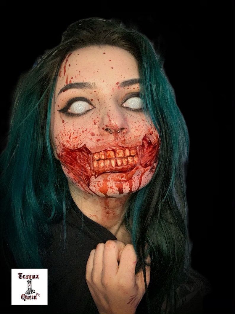 May include: A person with teal hair and white contact lenses featuring special effects makeup that simulates a torn mouth with exposed teeth and blood splatters across the face. A small logo in the corner displays the text Trauma Queen.