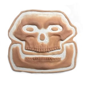 Peut inclure: Une pièce prothétique en silicone plate, beige et blanche, en forme de crâne humain avec une mâchoire inférieure séparée. Le design présente des détails anatomiques en relief couleur peau, incluant orbites, cavité nasale et dents.