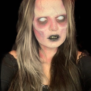 May include: A person with long brown hair featuring blonde highlights, wearing black clothing. The face is painted with pale white foundation, dark red contouring around the eyes and forehead, and black lipstick, with solid white contact lenses.