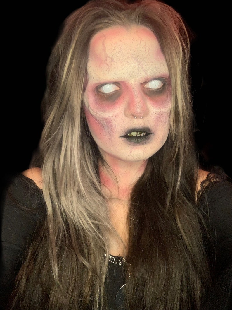 May include: A person with long brown hair featuring blonde highlights, wearing black clothing. The face is painted with pale white foundation, dark red contouring around the eyes and forehead, and black lipstick, with solid white contact lenses.