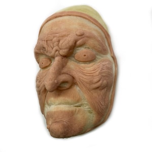 May include: A detailed, aged face mask with a wrinkled, textured surface. The mask features a prominent nose, sunken eyes, and a slightly open mouth. The color palette includes shades of tan, brown, and a hint of green, suggesting an old age theme.