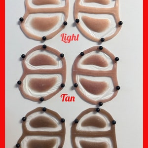 May include: Six silicone prosthetic eye patches arranged in three rows labeled Light, Tan, and Dark. Each patch features a translucent frame with two oval-shaped skin-toned inserts, secured by small black pins around the perimeter of the frames.