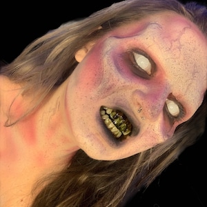 May include: A person with elaborate makeup featuring a mottled purple and pink complexion, white mesh contact lenses, and dark, stained teeth. The face has a textured, weathered appearance with a nose ring and long, light brown hair.