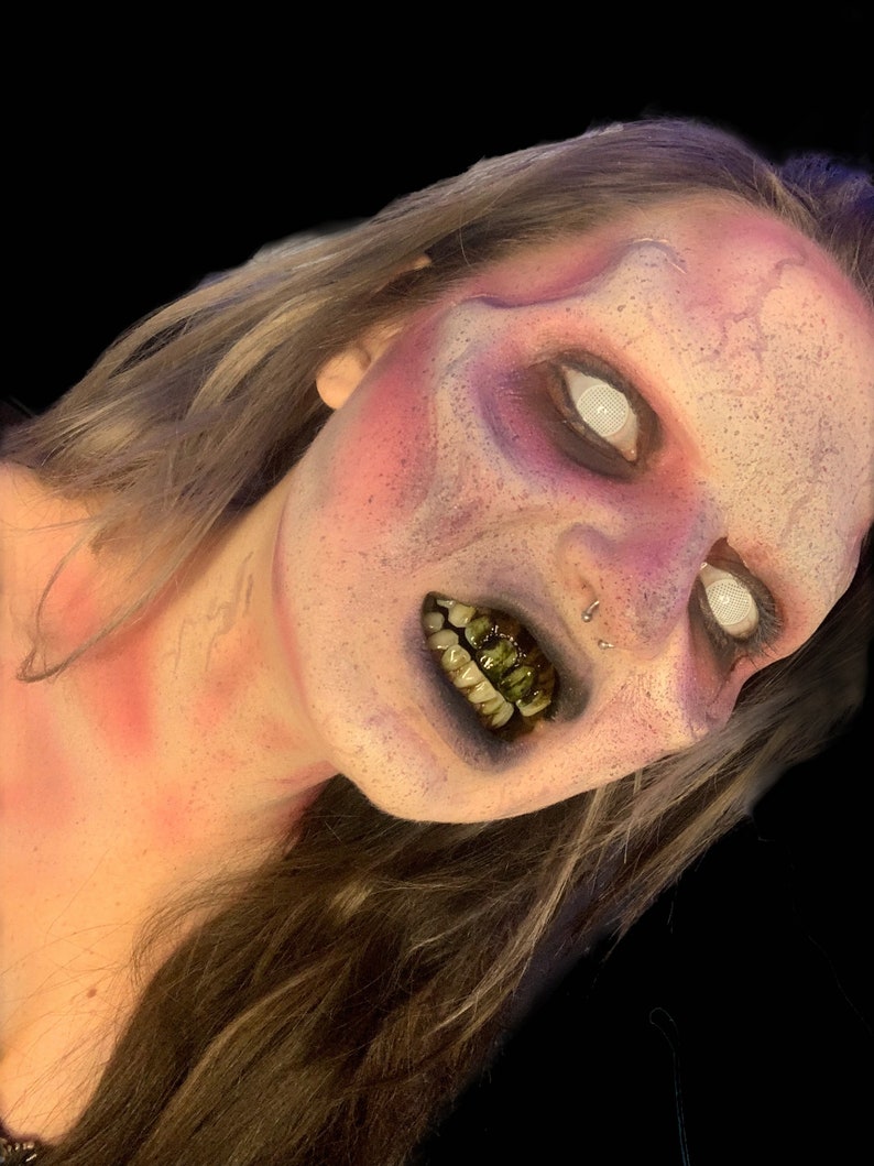May include: A person with elaborate makeup featuring a mottled purple and pink complexion, white mesh contact lenses, and dark, stained teeth. The face has a textured, weathered appearance with a nose ring and long, light brown hair.