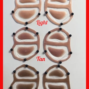 May include: Six silicone prosthetic eye patches arranged in three rows labeled Light, Tan, and Dark. Each patch features a translucent frame with two oval-shaped skin-toned inserts, secured by small black pins around the perimeter of the frames.
