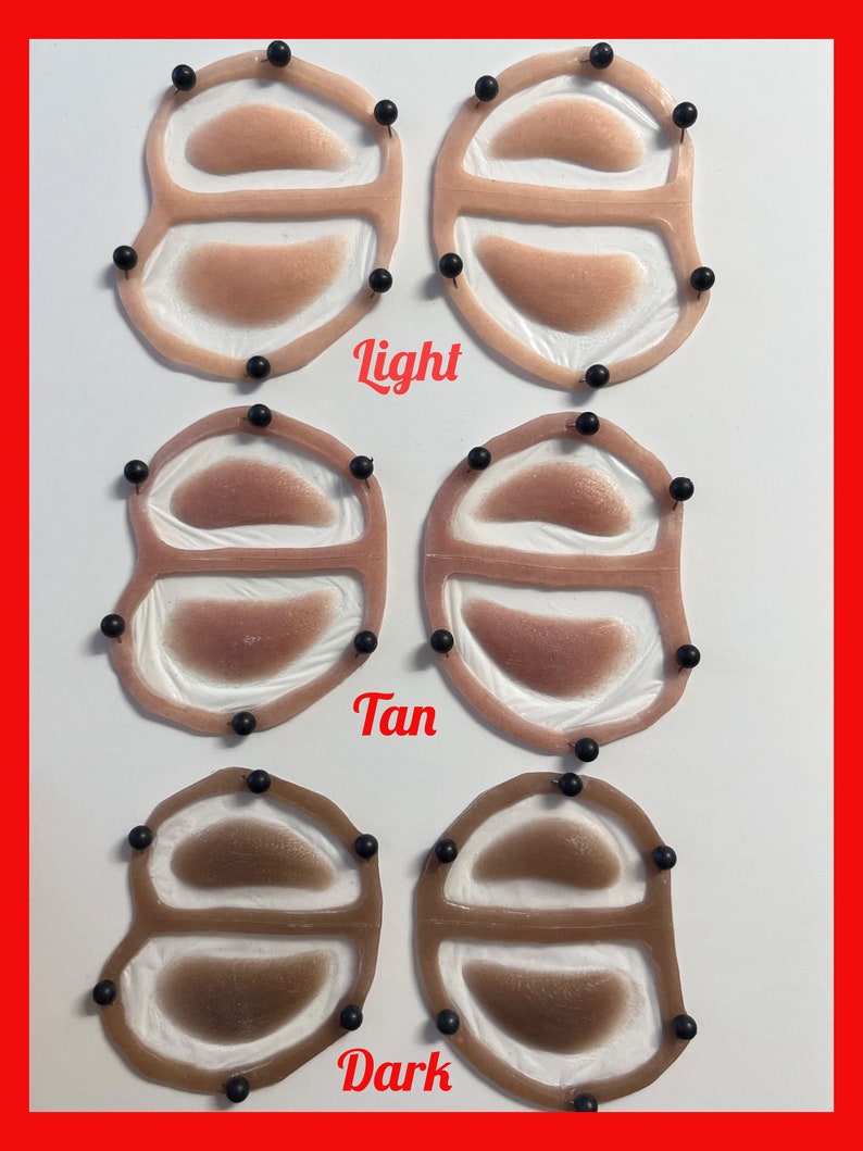 May include: Six silicone prosthetic eye patches arranged in three rows labeled Light, Tan, and Dark. Each patch features a translucent frame with two oval-shaped skin-toned inserts, secured by small black pins around the perimeter of the frames.