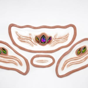 May include: Four white silicone molds with raised lines and a brown border. Each mold has a different shape and features iridescent green and purple rhinestones.