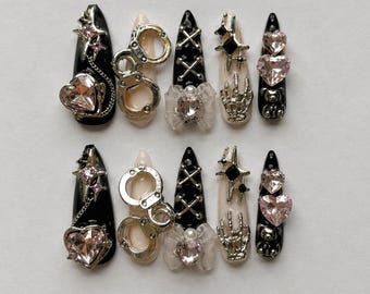May include: A set of ten press-on nails in black and nude tones, featuring silver-tone metal charms including handcuffs, skeleton hands, and cross-lacing. The nails are decorated with pink heart-shaped rhinestones, star accents, and bows.