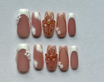 May include: A set of ten press-on nails featuring a nude base with white French tips, small white floral accents, and gold wire-wrapped flower designs. The nails have a square-tapered shape and include abstract white cloud-like patterns.
