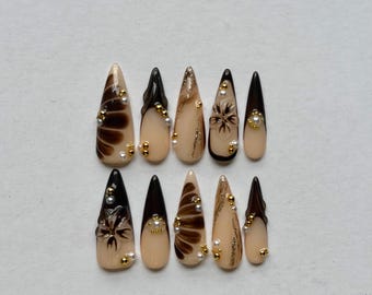 May include: A set of ten stiletto-shaped press-on nails featuring a nude base with black and brown abstract floral patterns, dark tips, and gold and white bead embellishments arranged in various designs across the individual nail surfaces.
