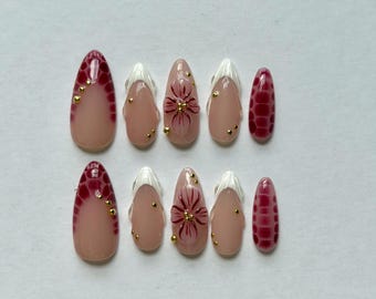 May include: A set of ten almond-shaped press-on nails featuring a nude base. Designs include deep red crocodile skin patterns, white French tips, and hand-painted dark red floral motifs accented with small, metallic gold spherical studs.
