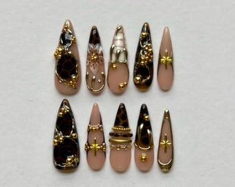 May include: A set of ten stiletto-shaped press-on nails featuring a nude base with brown leopard print patterns, gold metallic accents, small gold beads, and star-shaped charms. The designs include intricate gold wire detailing and glossy finishes.