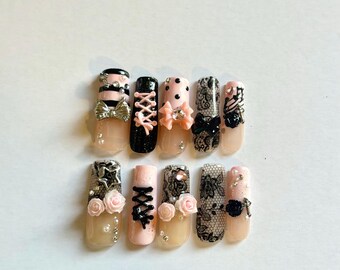 May include: A set of ten press-on nails featuring a color palette of soft pink, black, and white. The designs include black lace patterns, pink rose embellishments, silver bows, corset-style lacing, polka dots, stripes, and small rhinestones.