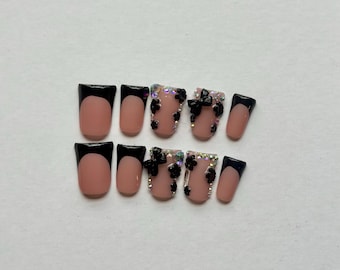 May include: A set of ten press-on nails featuring a nude base with black French tips. Some nails are adorned with black floral embellishments and clear iridescent rhinestones, while others maintain a simple black and nude French tip design.