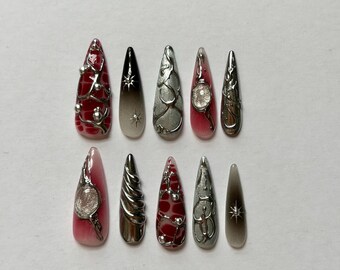 May include: A set of ten stiletto-shaped press-on nails featuring a mix of designs. Some nails have a deep red base with raised silver metallic liquid-like accents, while others feature black-to-clear gradients, silver star charms, and textured silver swirls.