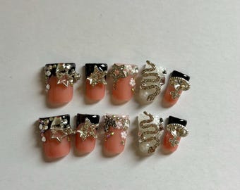 May include: A set of ten press-on nails featuring a peach base with black tips. The designs include gold star charms, small white floral accents, clear rhinestones, a gold snake-shaped embellishment, and a gold circular charm with a cross detail.