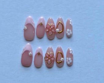 May include: A set of ten almond-shaped press-on nails in a soft pink base color. The designs include white floral accents, gold starbursts, raised pink flower petals with gold outlines, and gold abstract shapes, all finished with a glossy top coat.