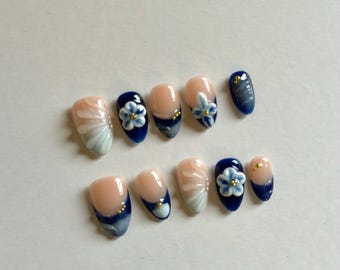 May include: A set of ten press-on nails featuring a glossy finish with a nude base and deep navy blue tips. The design includes hand-painted white floral accents, swirling blue marble patterns, and small gold metallic studs on several of the nails.
