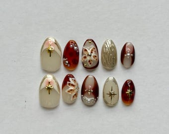 May include: A set of ten press-on nails featuring a color palette of cream, deep red, and gold. The designs include gold star charms, raised floral accents, metallic gold textures, and small white pearls arranged on various oval-shaped nail tips.