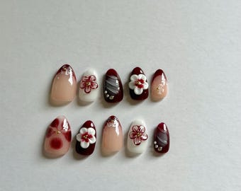 May include: A set of ten almond-shaped press-on nails featuring a color palette of deep burgundy, white, and nude. The designs include 3D white floral accents with red centers, gold metallic studs, and hand-painted red floral patterns on solid bases.