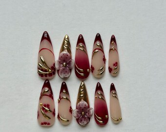 May include: A set of ten almond-shaped press-on nails featuring a nude base with deep burgundy tips. The design includes gold metallic accents, delicate hand-painted floral patterns, and three-dimensional flower embellishments in a soft purple.